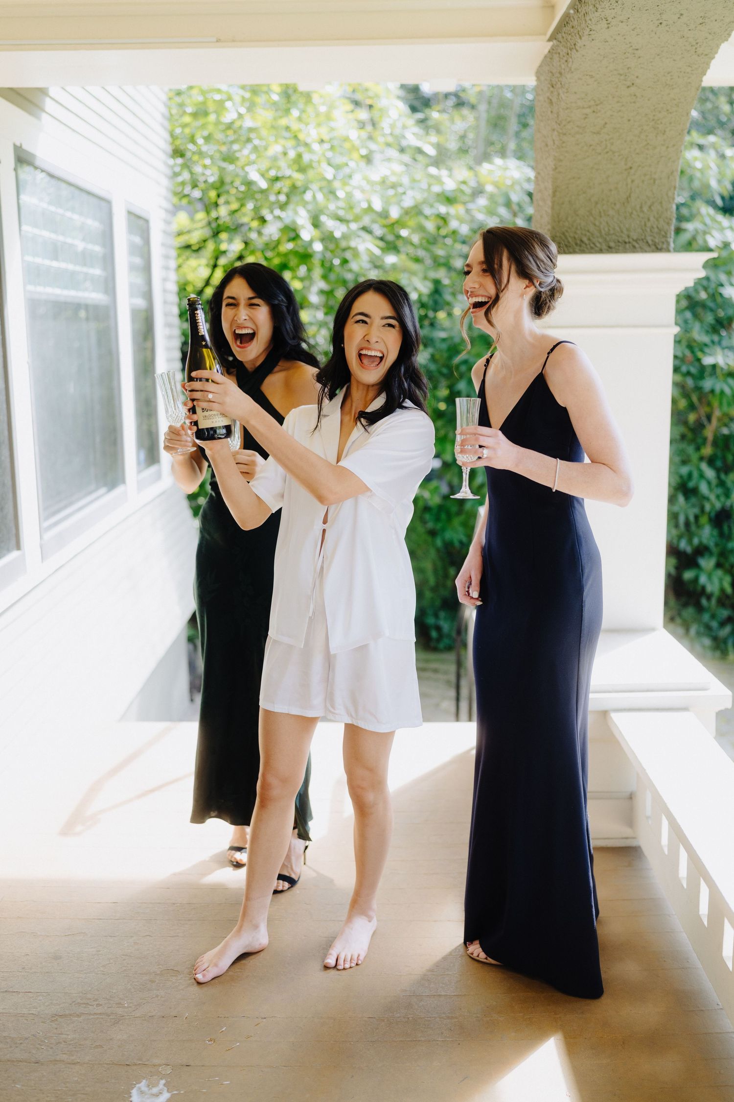 Three friends in formal attire celebrating with drinks and laughter on a sunny porch.