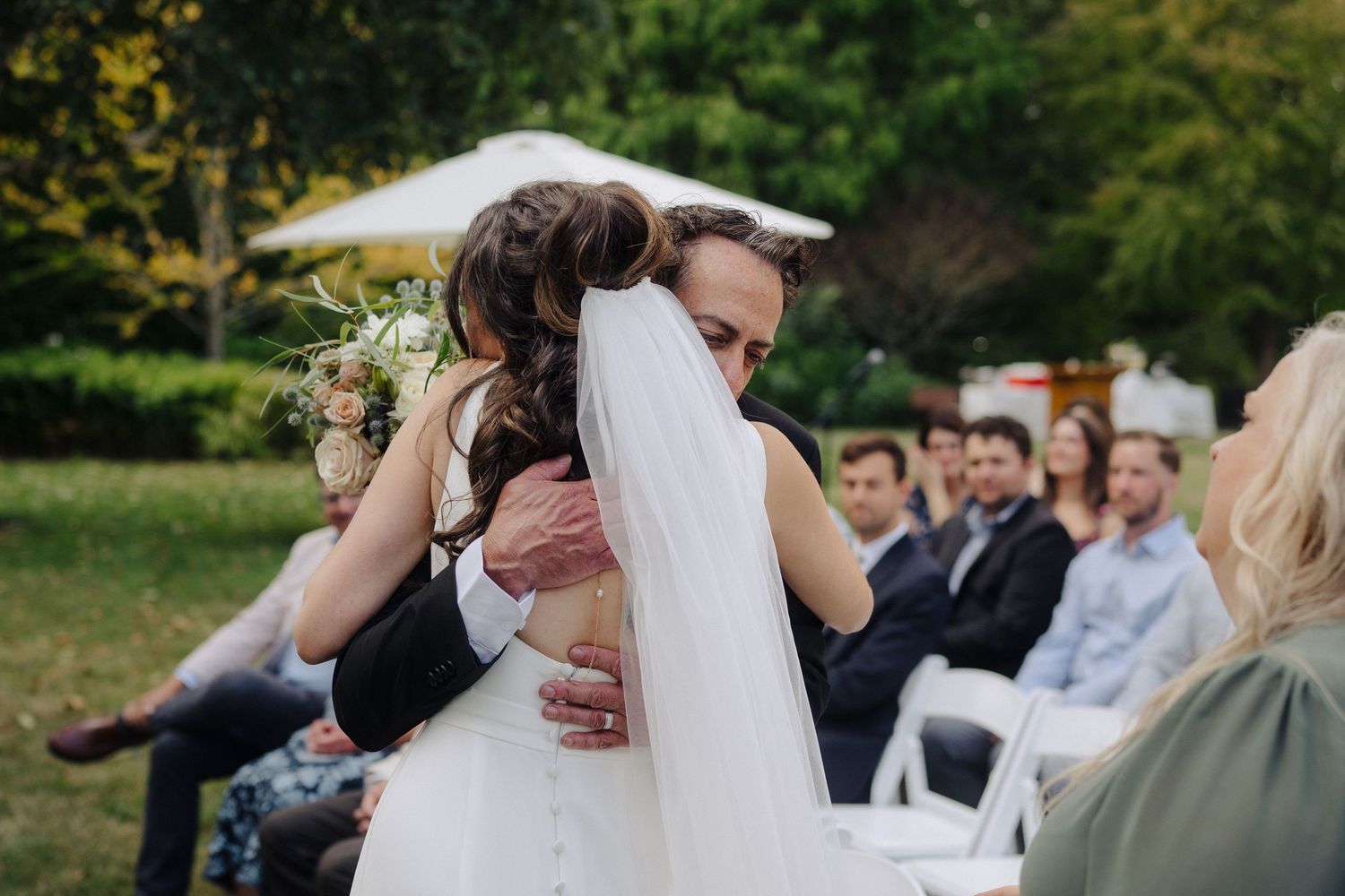 Emotional embrace during outdoor wedding ceremony with guests seated in background.
