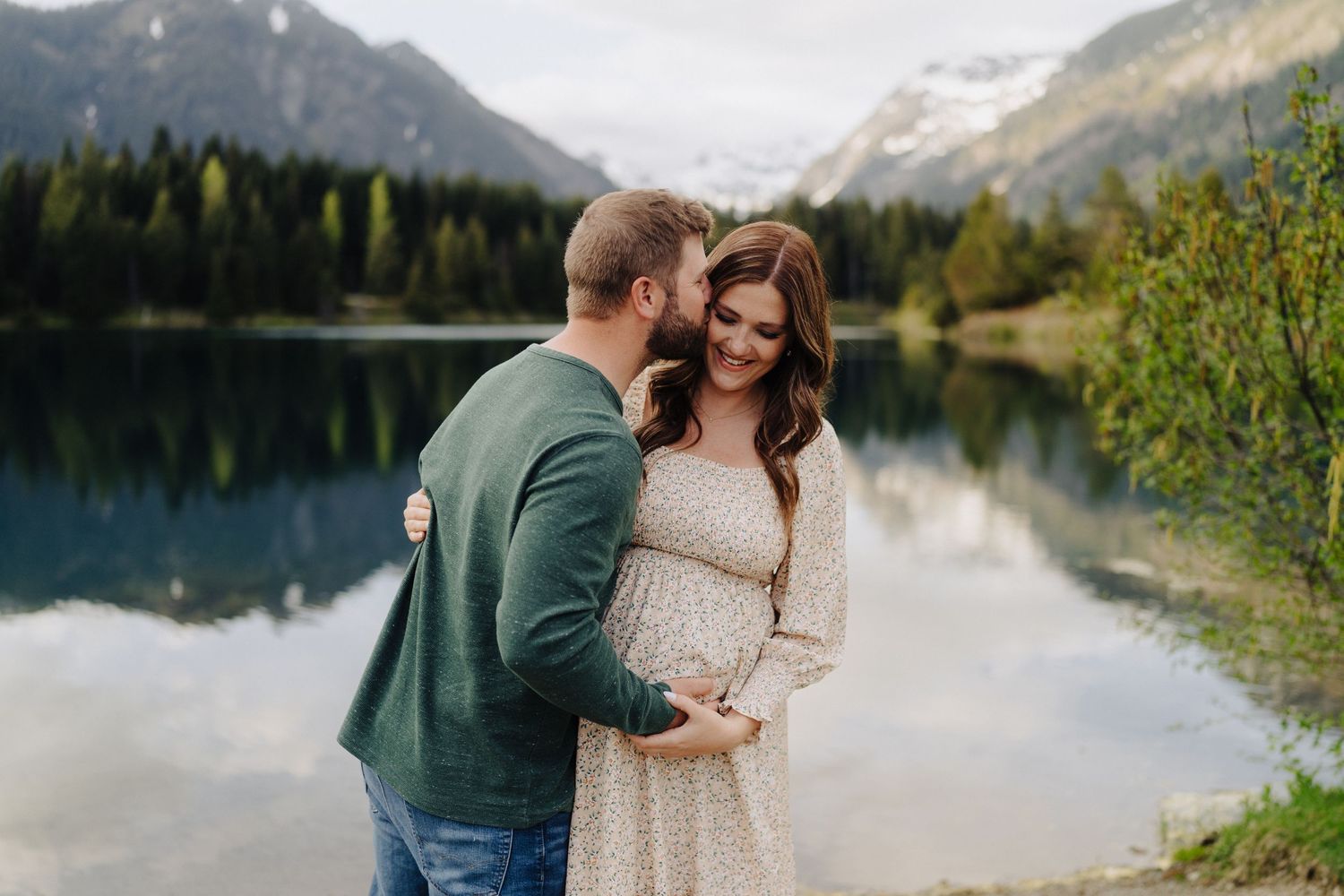 Couple embraces by a serene mountain lake surrounded by evergreen forest and snow-capped peaks at sunset.