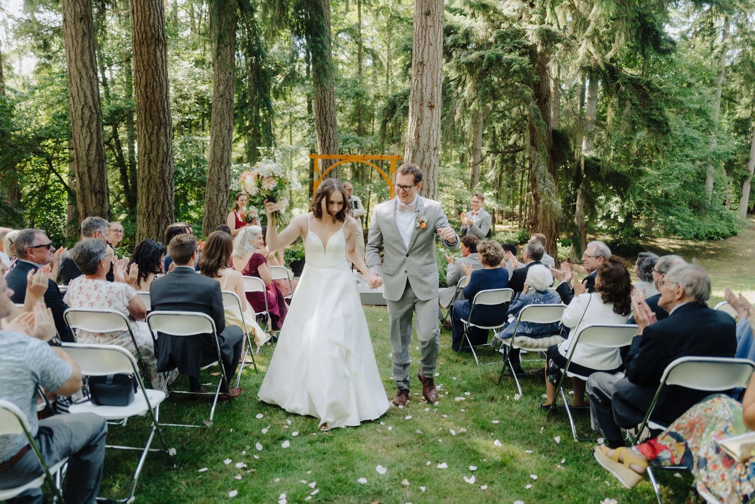 Outdoor wedding ceremony in progress among tall trees with guests seated on white chairs.