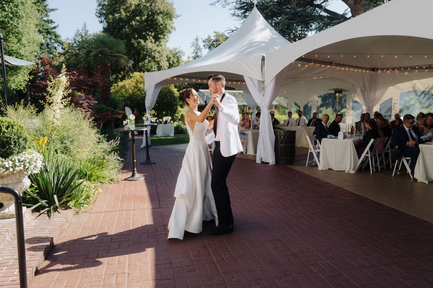 Wedding reception setup under a white tent with brick pathways and garden surroundings.