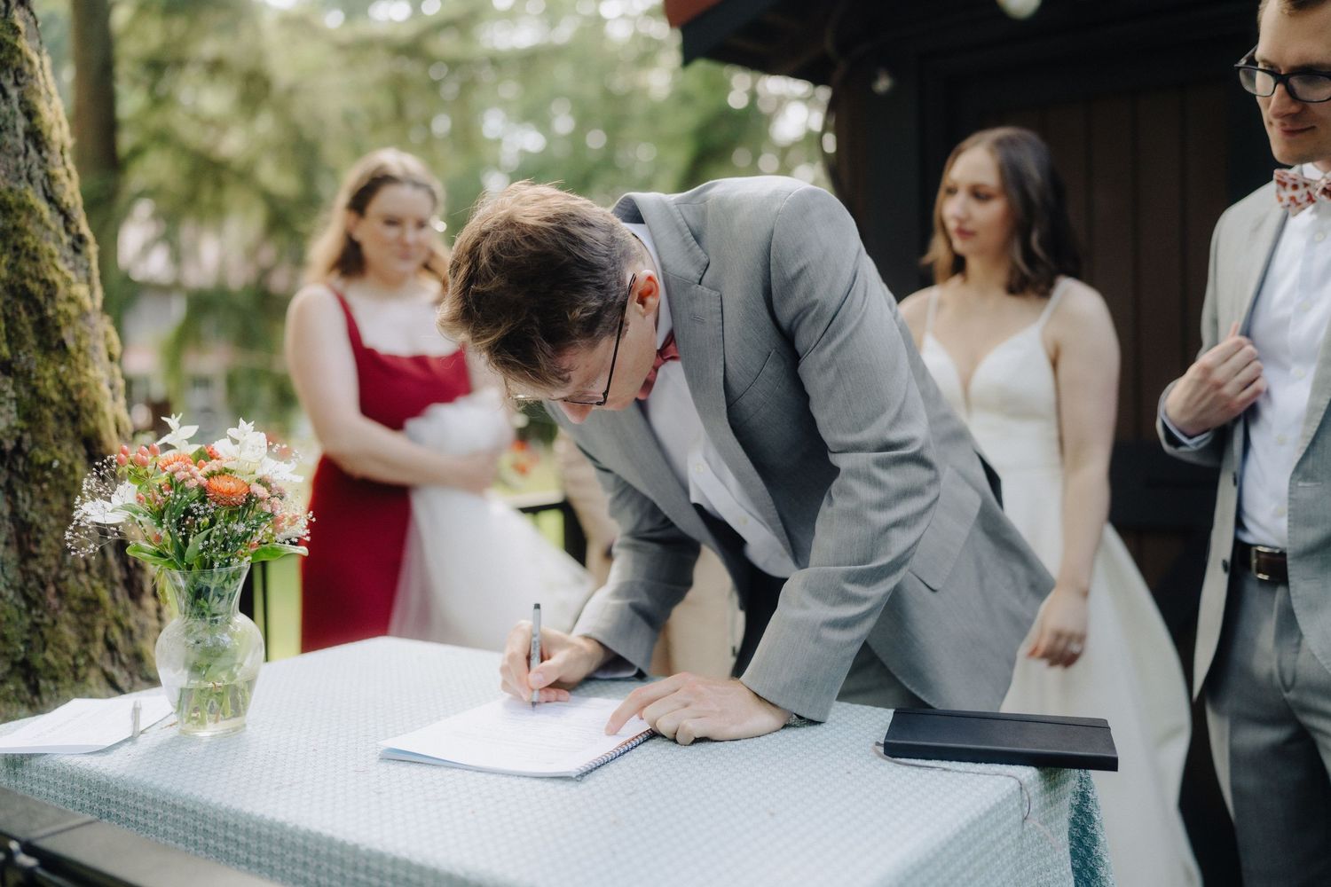 Couple signing marriage documents at an outdoor wedding ceremony with witnesses gathered around.