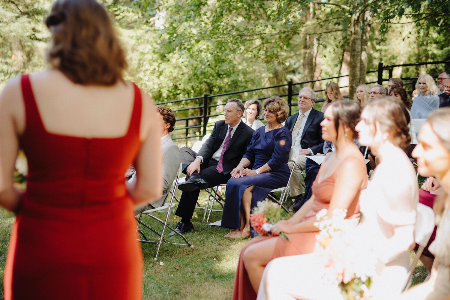Wedding ceremony guests watching from chairs with bridesmaids in red dresses visible.