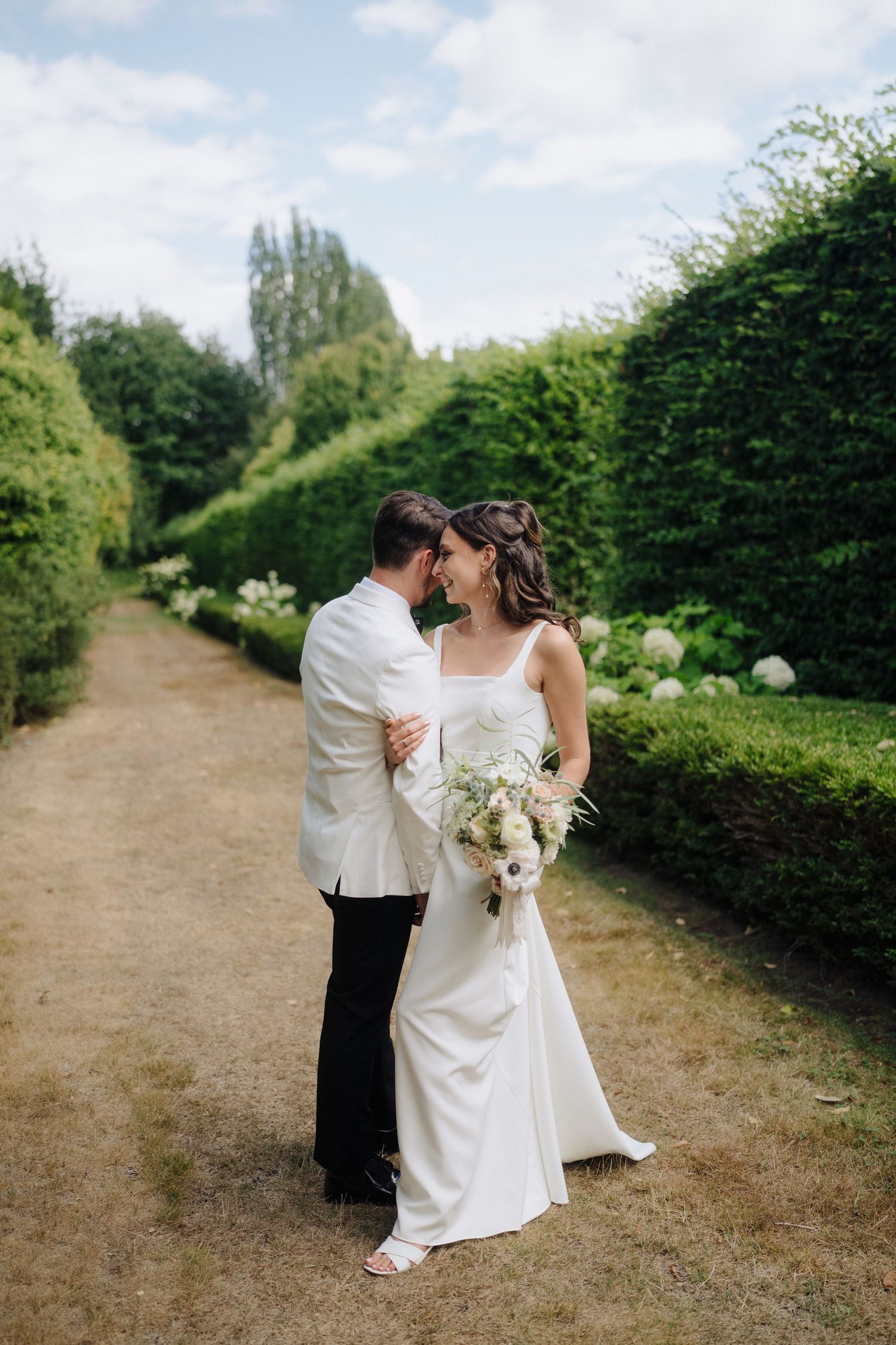 Romantic wedding couple poses along garden path lined with tall green hedges.