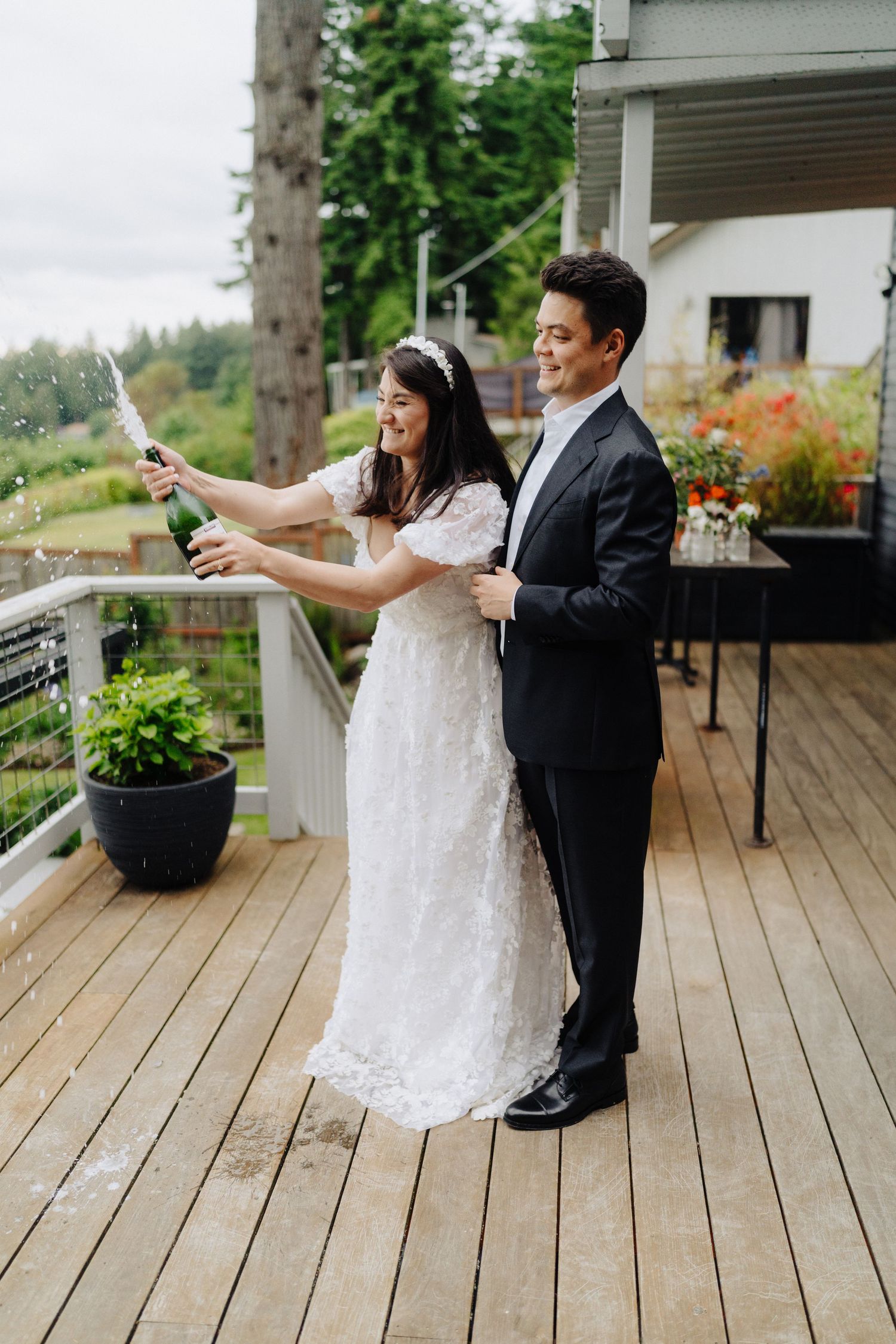 A couple pops champagne together on their wedding day while standing on a deck with mountain views.