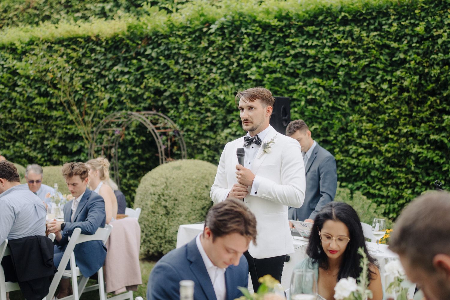 Guests seated at outdoor wedding reception tables surrounded by lush green hedges.