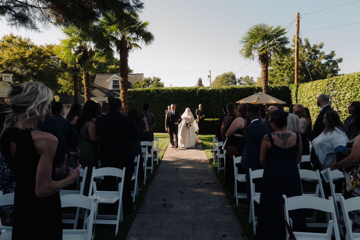 Wedding ceremony in progress with guests seated in white chairs along a garden pathway.