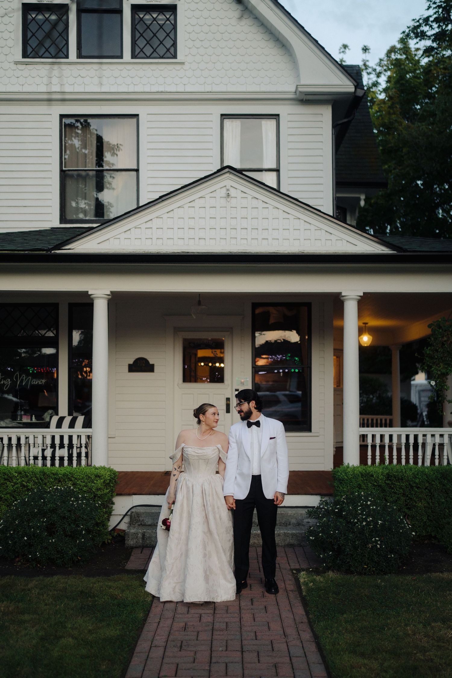 A couple in wedding attire stands on a brick pathway outside a white house at dusk.