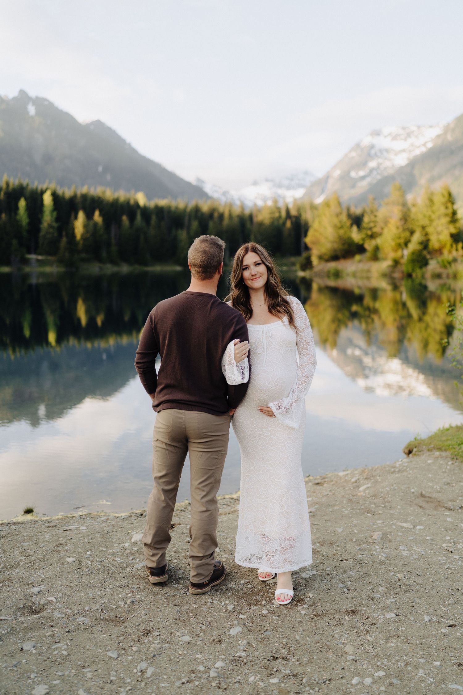 A couple stands by a serene mountain lake at sunset, surrounded by evergreen trees and snow-capped peaks in the distance.