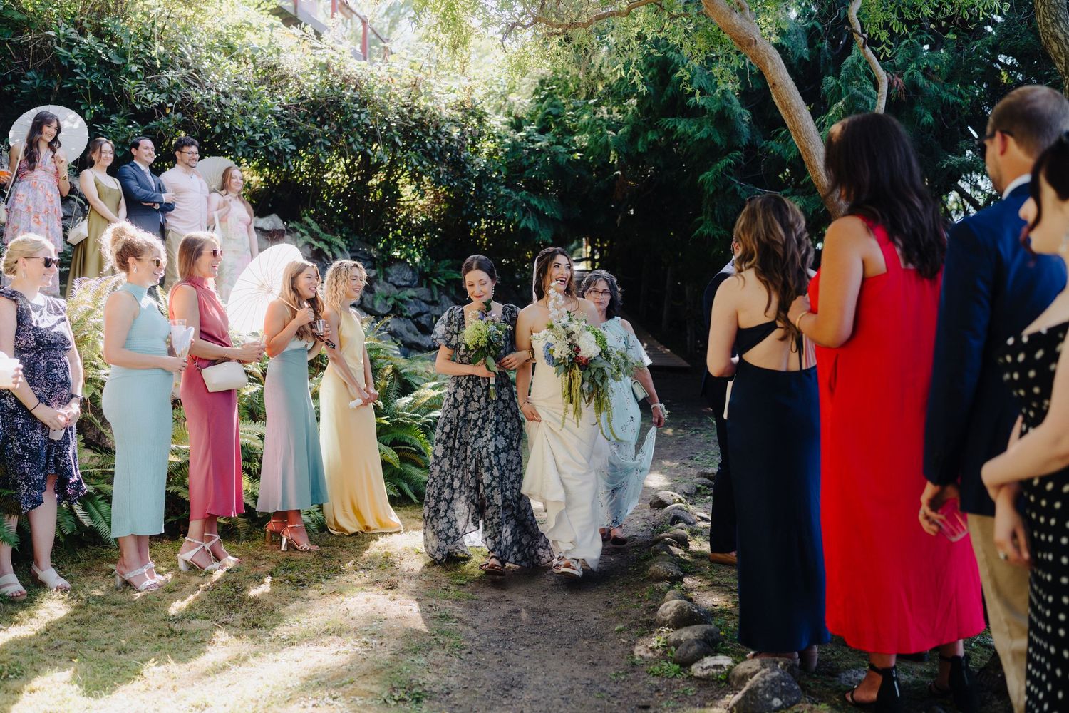 Wedding party and guests mingle on a garden path during an outdoor celebration.