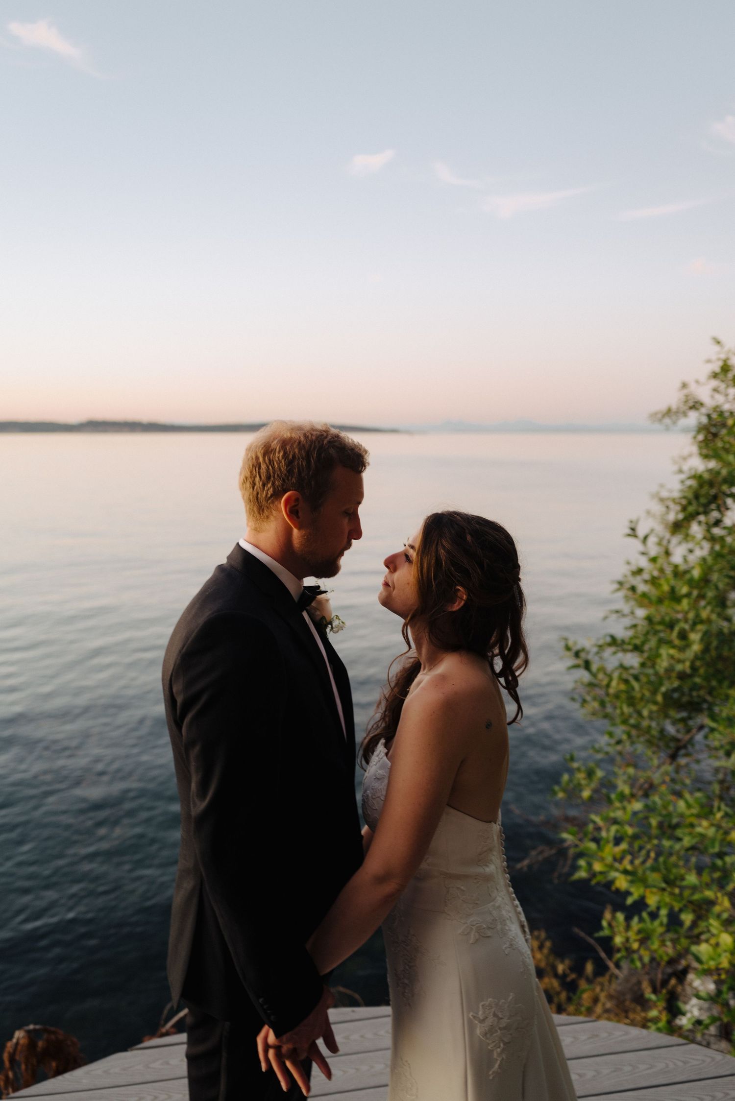 Newlyweds embrace on a lakeside deck during golden hour with water and trees in the background.