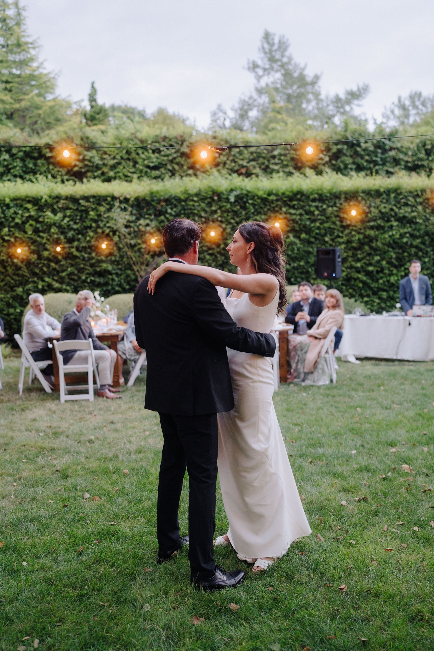 Couple dances beneath string lights at dusk during their outdoor garden reception.