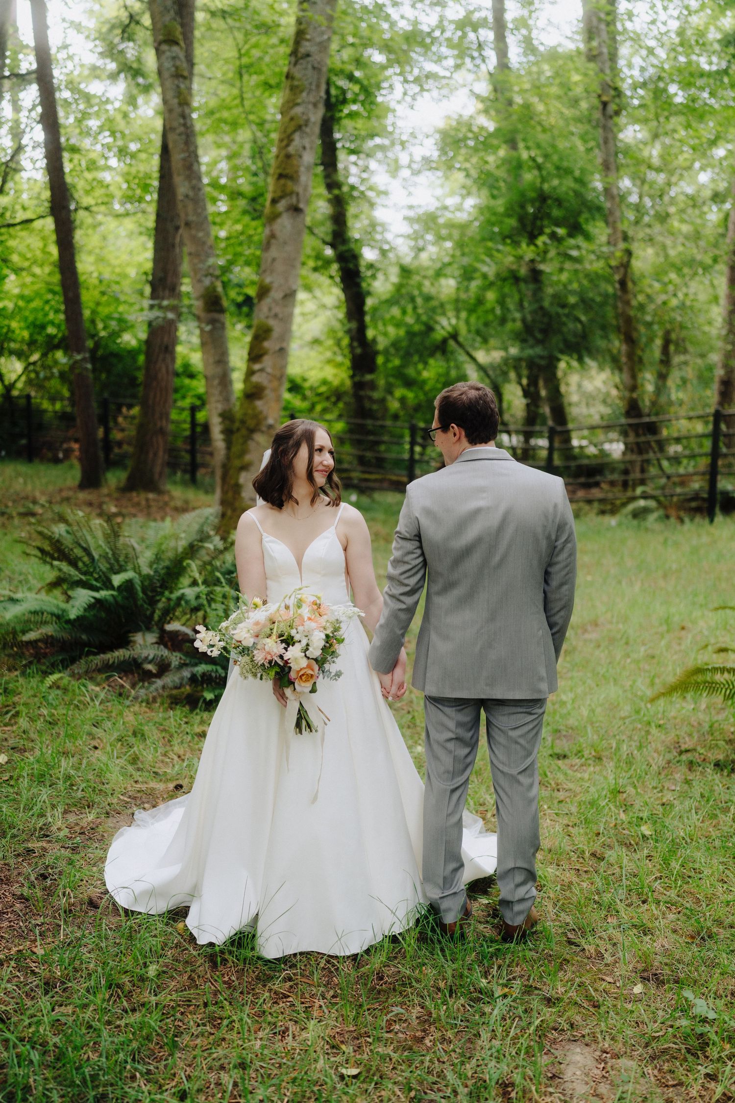A couple walks through a lush green wooded area in wedding attire, with the bride holding a beautiful floral bouquet.
