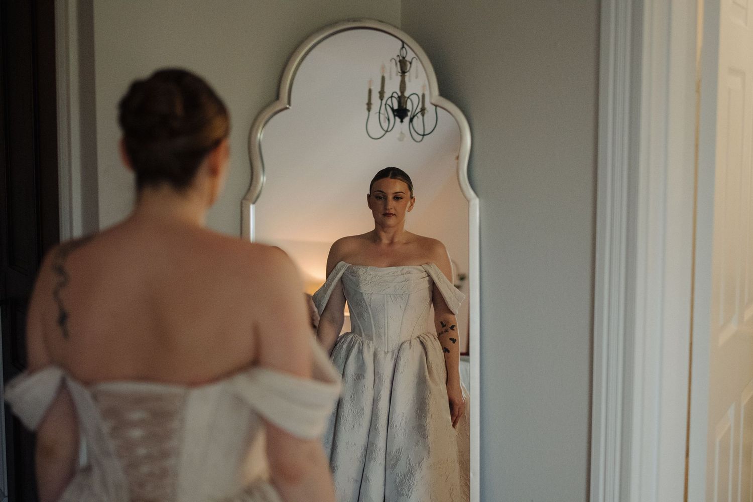 Person in a white dress stands in front of a mirror in an elegant room with chandelier lighting.