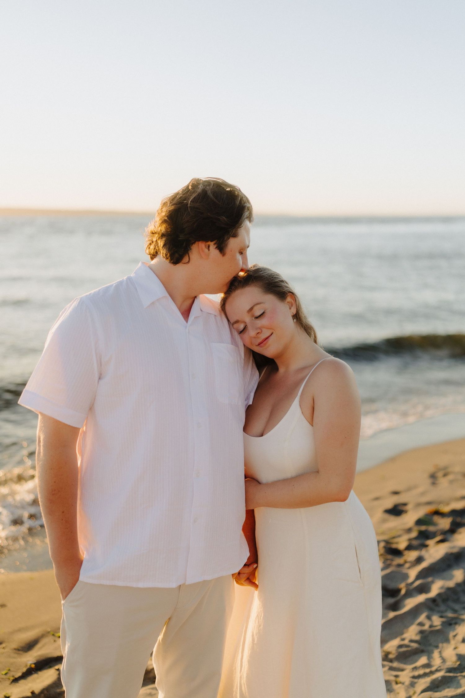 Romantic couple in white outfits embracing on a beach at sunset with ocean waves in the background.