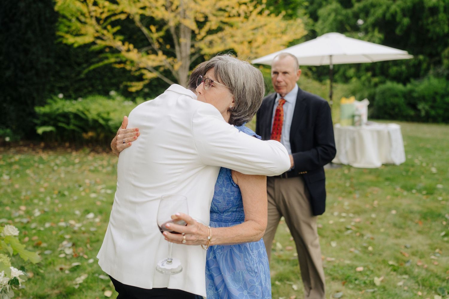 Wedding guests embrace during an outdoor garden celebration with white umbrellas in the background.