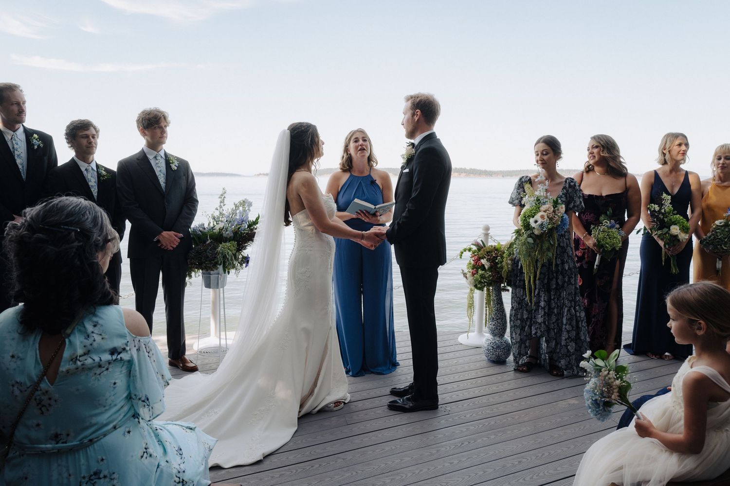 Wedding ceremony guests gather on a waterfront deck overlooking the ocean at sunset.