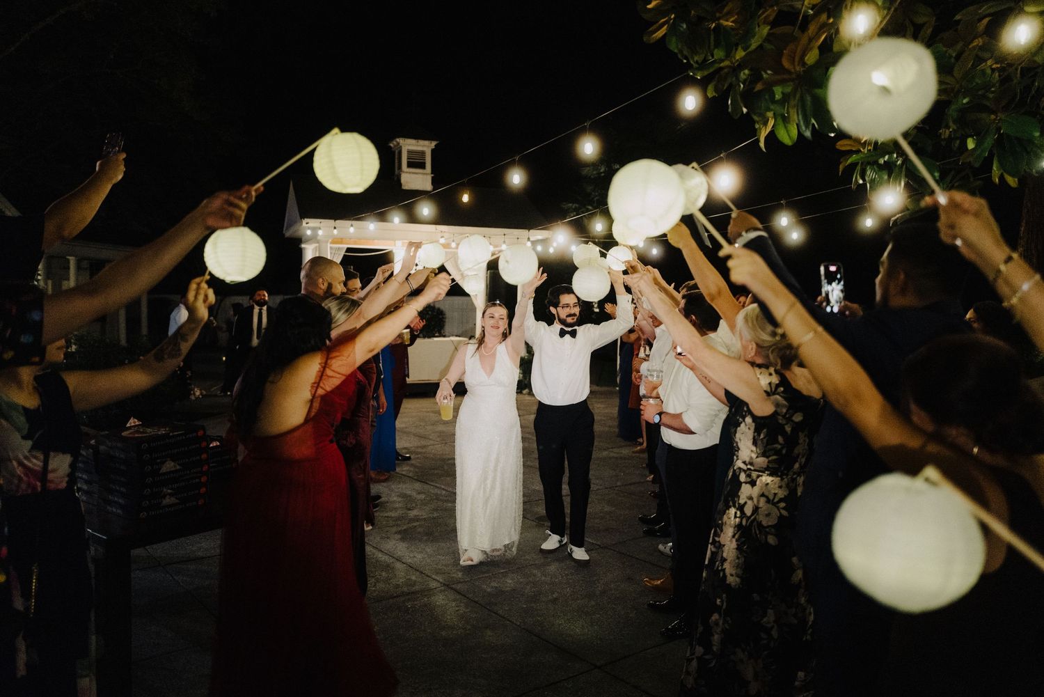 Wedding guests celebrate under glowing paper lanterns and string lights at an outdoor evening reception.