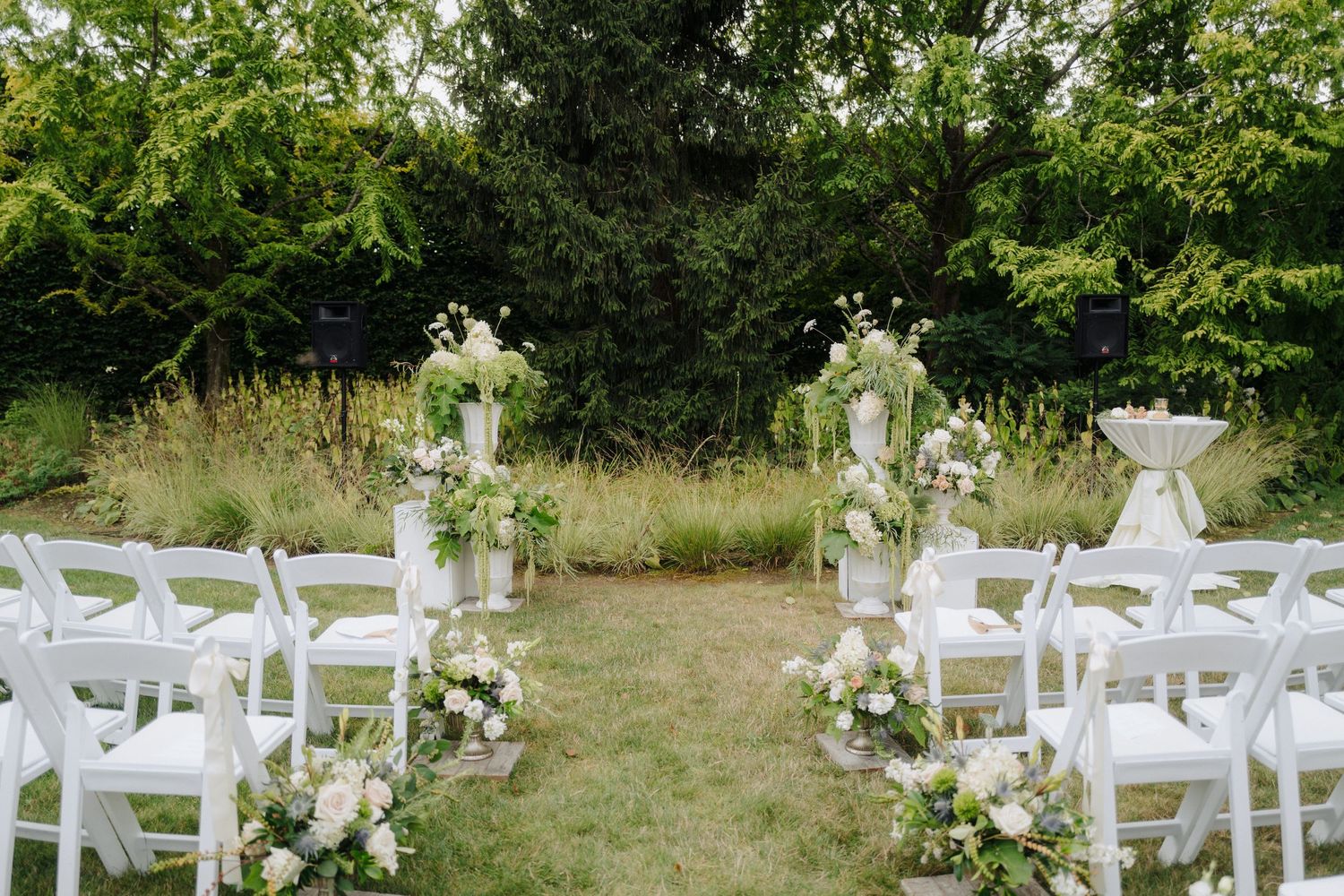 White chairs and floral arrangements set up for an outdoor garden wedding ceremony.