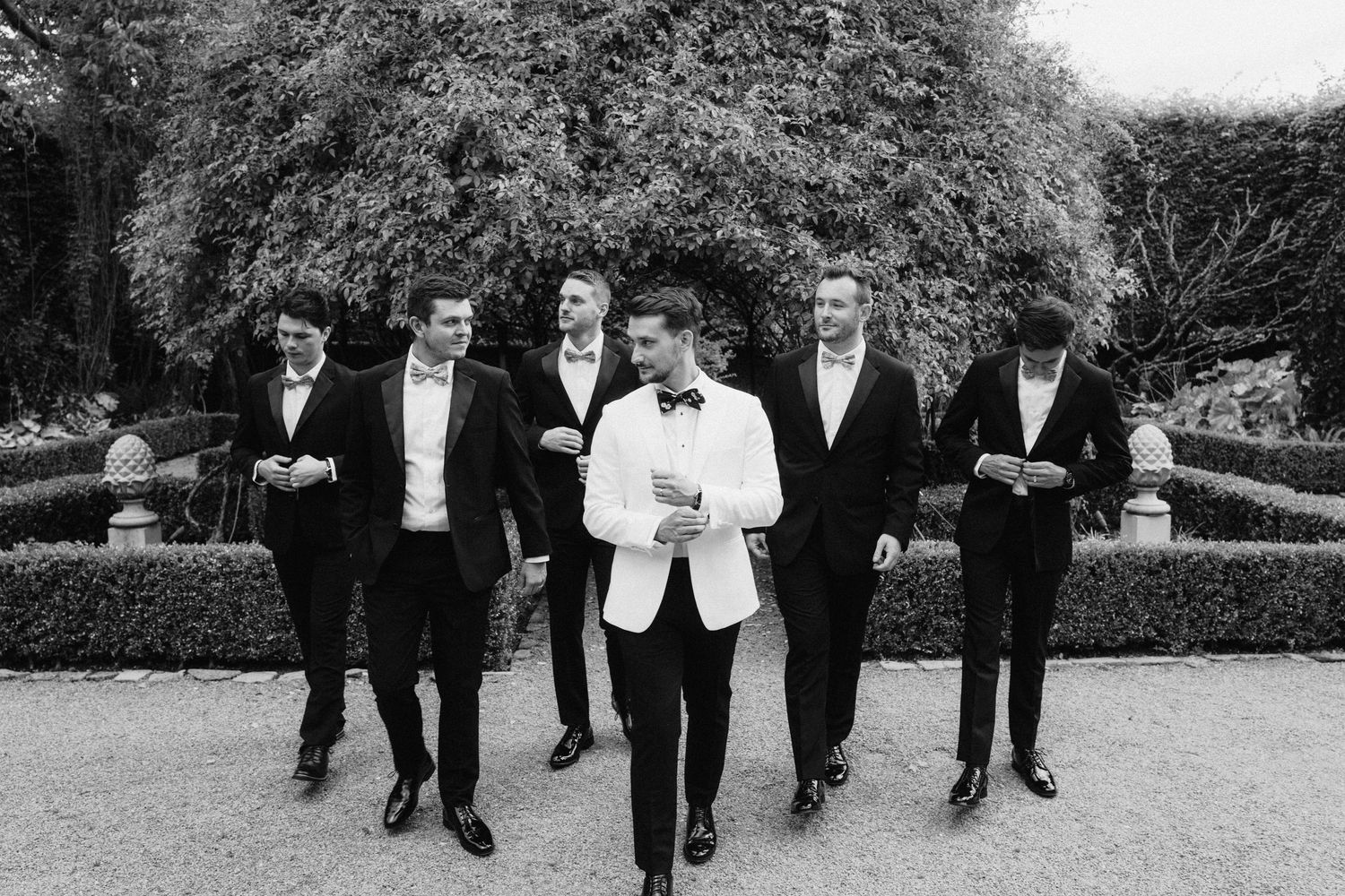 Groomsmen in black tuxedos walking and posing outdoors in a garden setting.