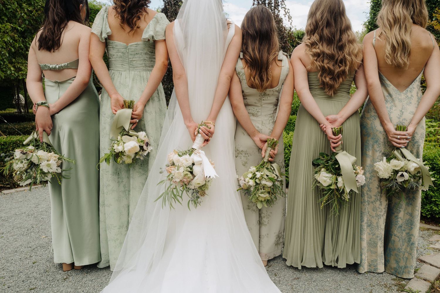 Bridesmaids in sage green dresses hold white floral bouquets while standing in a row.