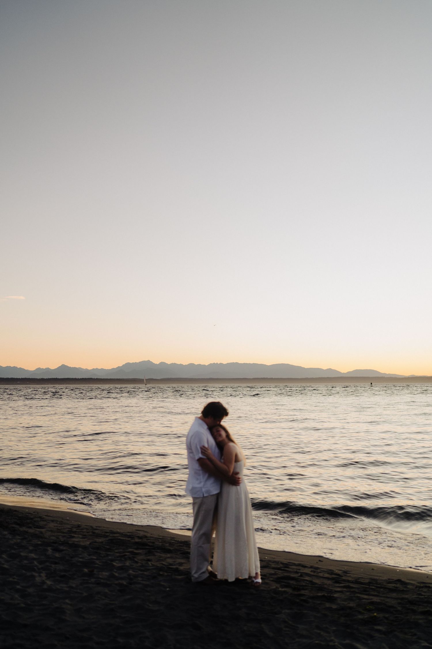 A couple embraces on a beach at sunset with mountains visible across the water and a golden sky above.