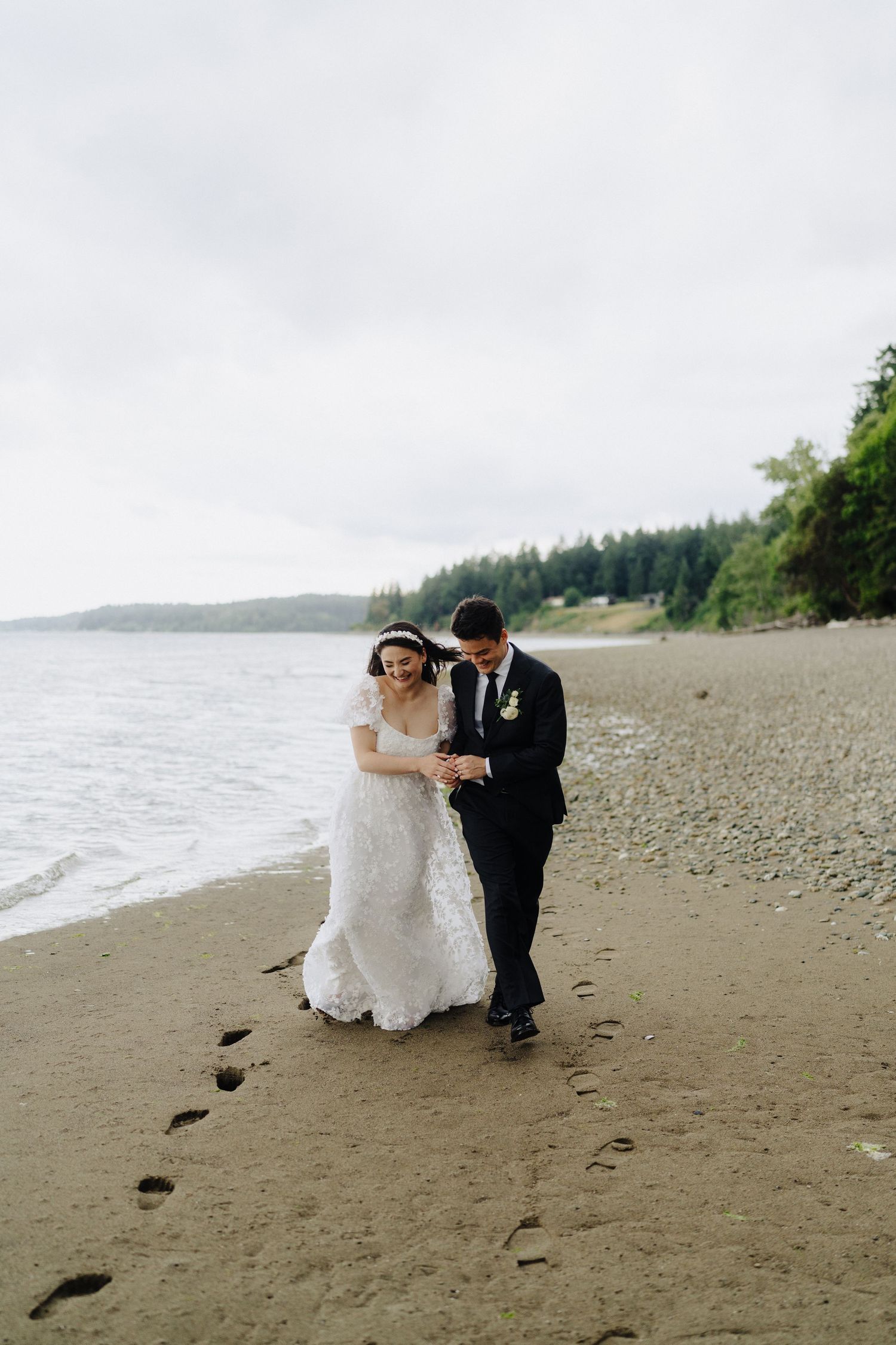 A couple walks together on a scenic beach leaving footprints in the sand with evergreen trees in the background.