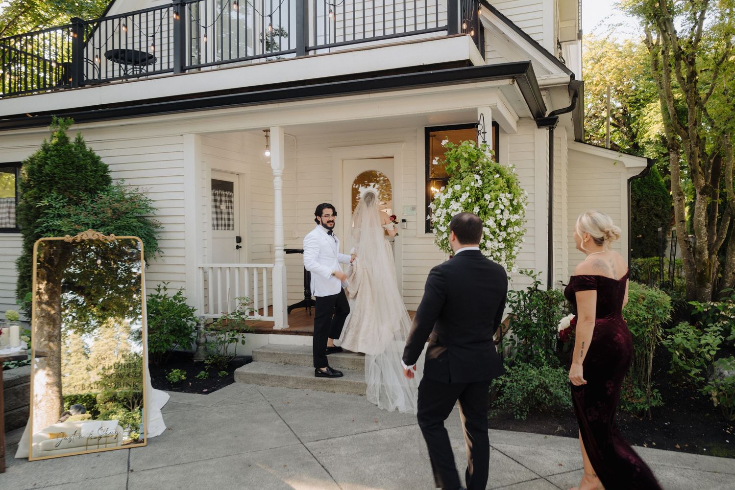Wedding party members gather on a white porch of a classic house during golden hour.