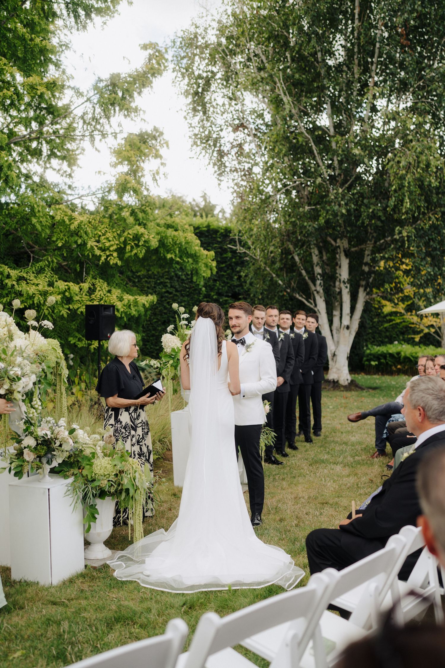 An outdoor wedding ceremony setup with white floral arrangements and guests gathered in a garden setting.