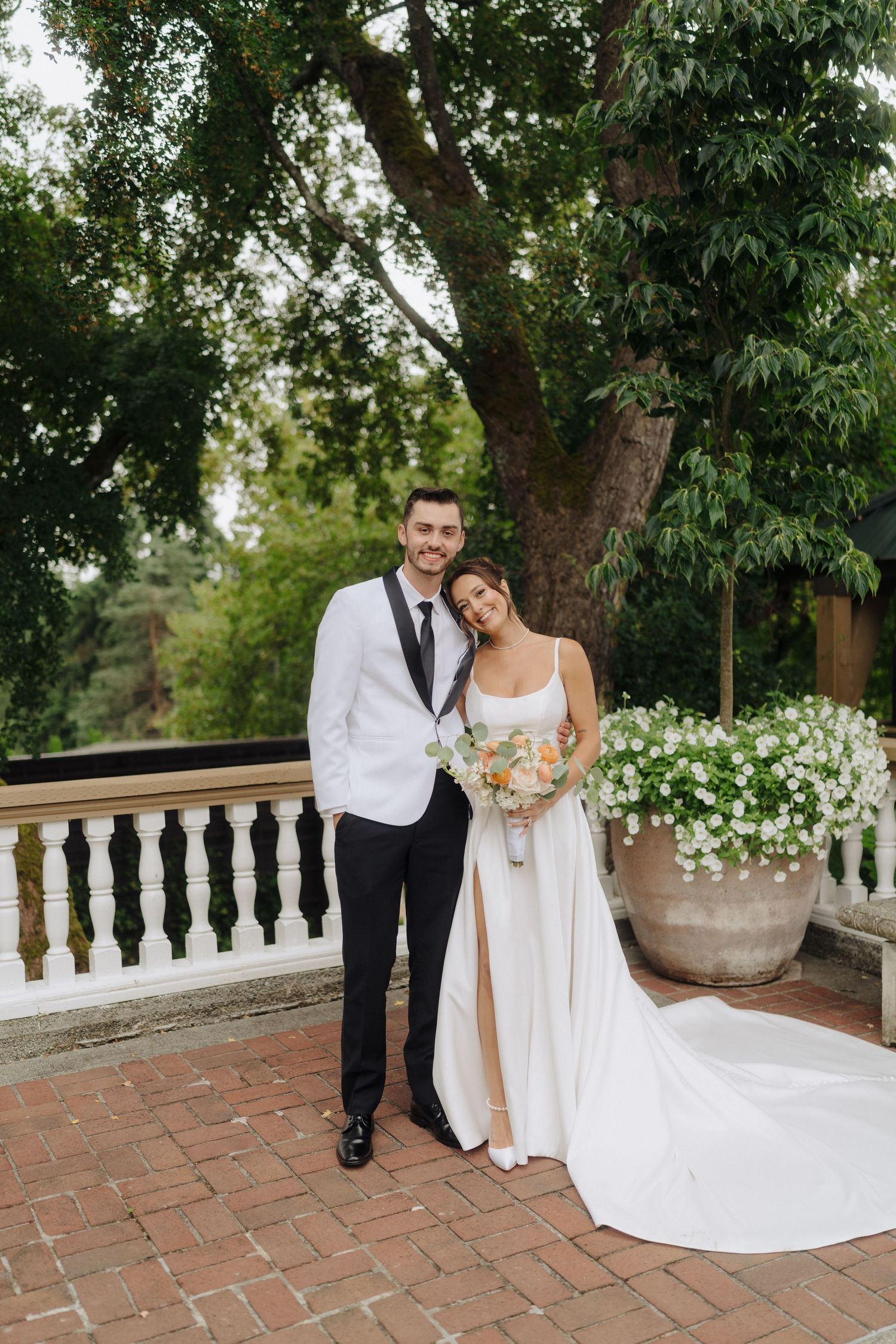 Newlywed couple poses on brick patio with stone balustrade and lush garden backdrop.