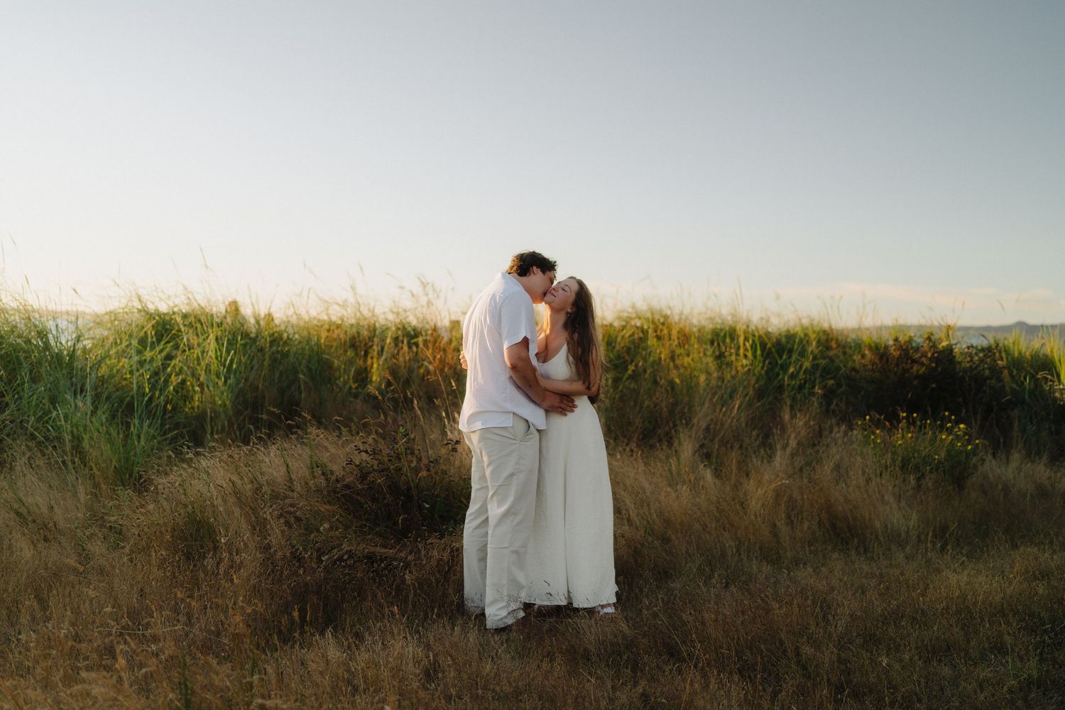 Romantic couple walking along a scenic beach path through tall grass at sunset.