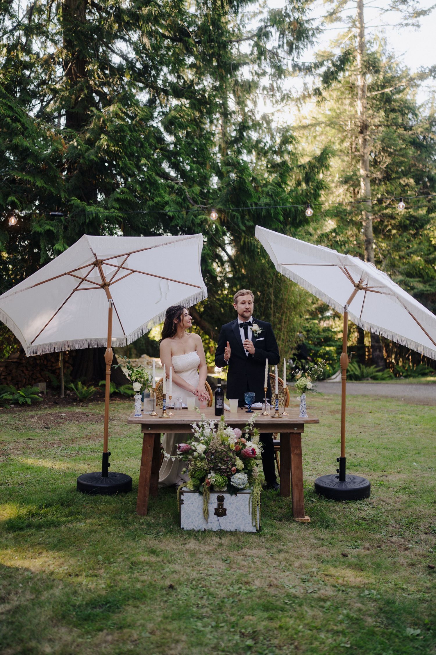 Outdoor wedding reception setup with white market umbrellas and wooden tables on grass.