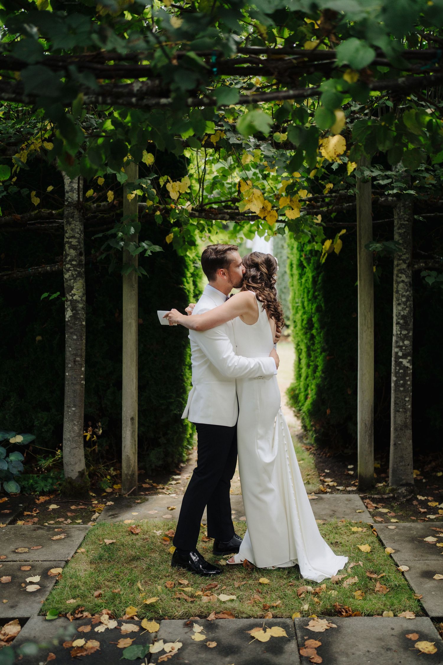 Wedding couple embracing under a canopy of trees with fallen autumn leaves scattered on the ground.