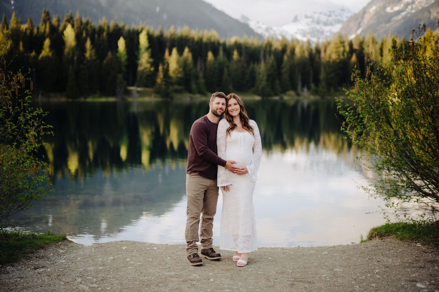 Couple poses by tranquil mountain lake surrounded by evergreen trees and snow-capped peaks at sunset.