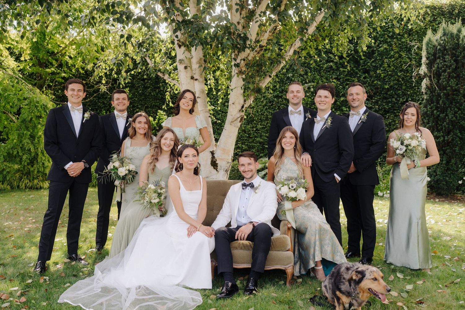 Wedding party poses for formal photos in an elegant garden setting with bridesmaids in sage green dresses.