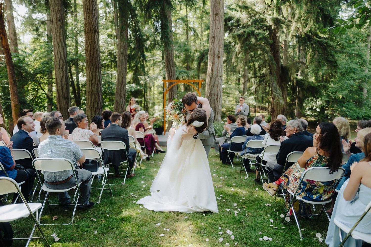 Wedding ceremony in progress at forest venue with white chairs and natural backdrop.