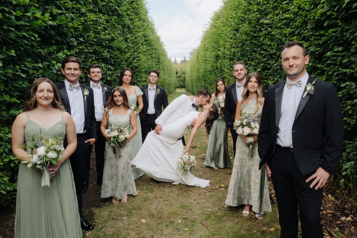 Wedding group poses in a beautiful garden alley with tall green hedges creating a natural corridor.