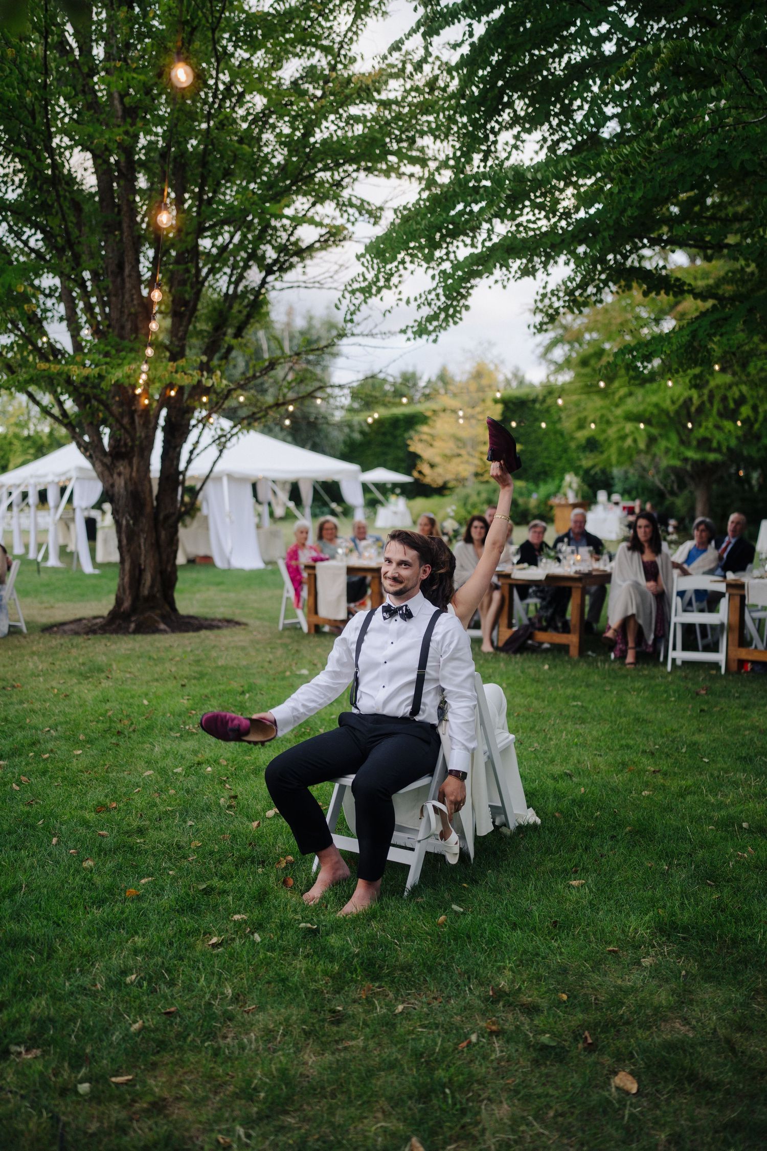 Guests enjoy an outdoor wedding reception under string lights and large shade trees.