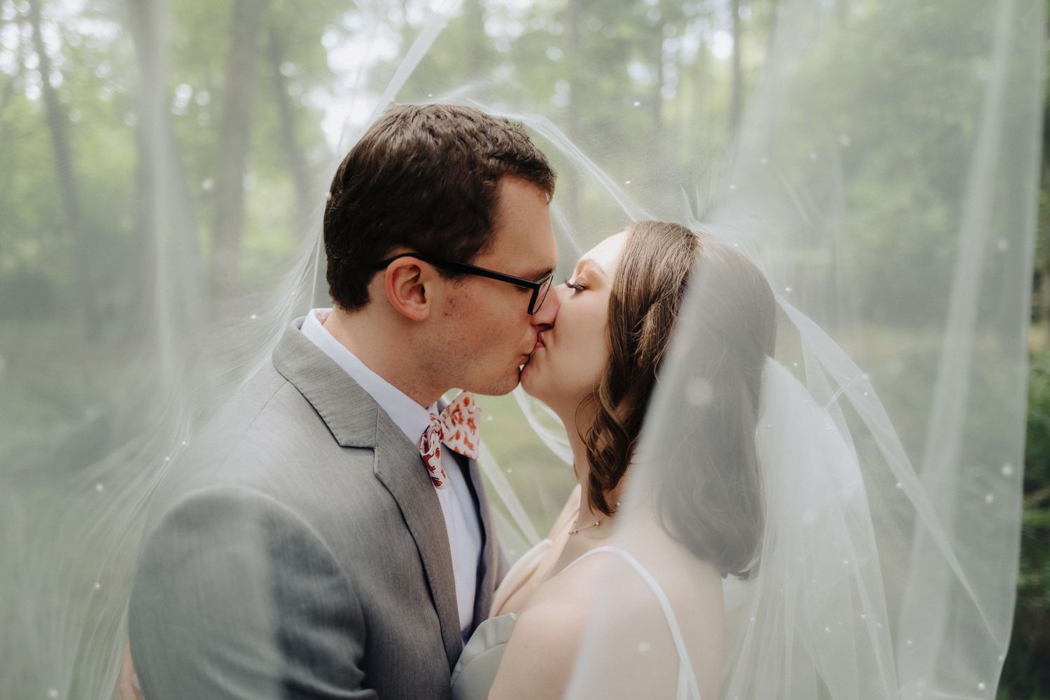 Romantic wedding kiss captured through flowing white veil in forest setting.