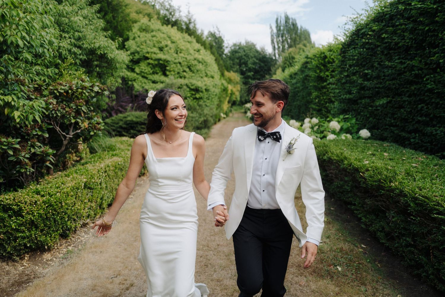 Newlyweds walk hand in hand through a garden path lined with lush hedges and greenery.