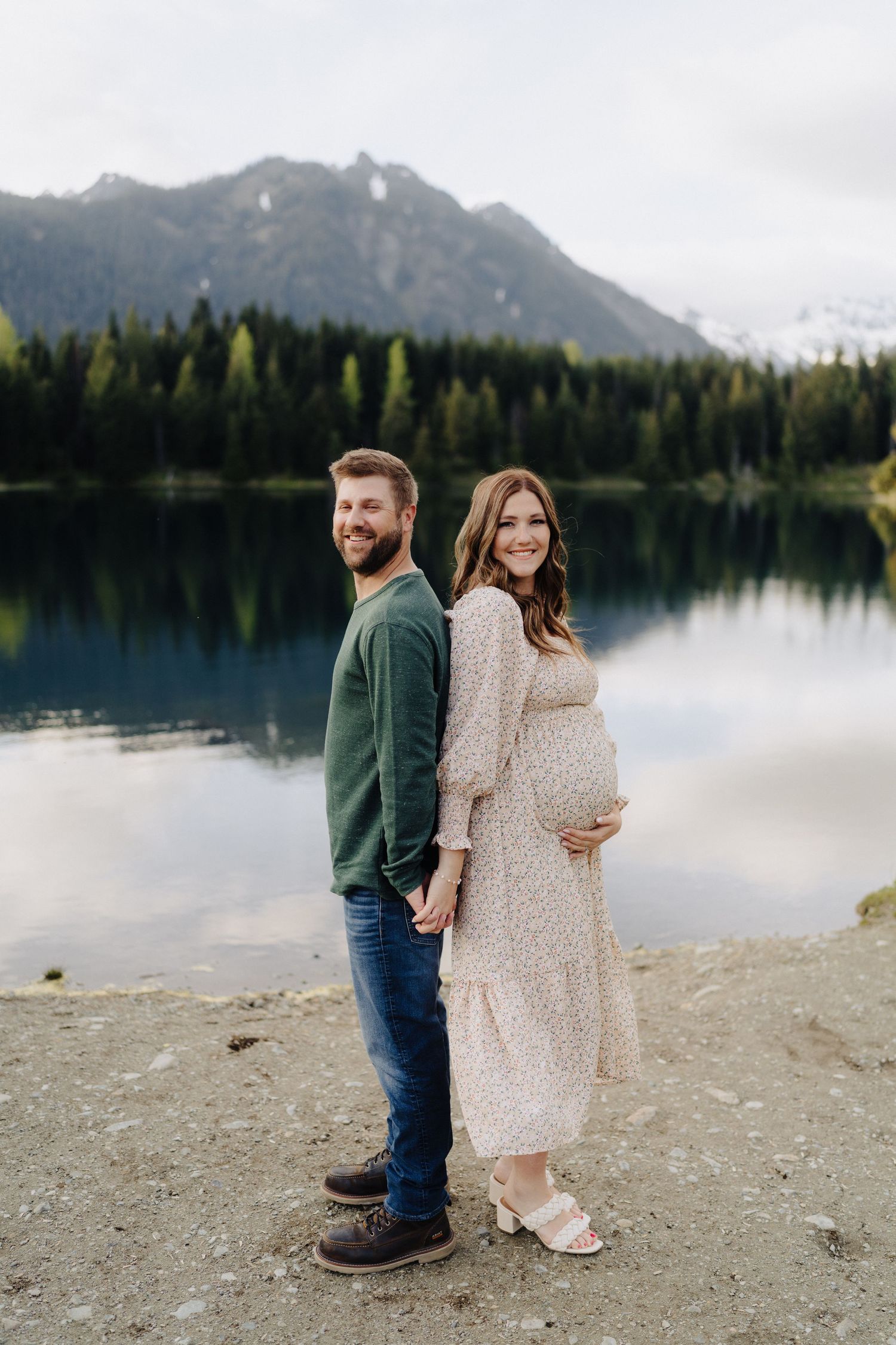 A couple poses together on a rocky beach with mountains and evergreen forest reflected in a calm lake.