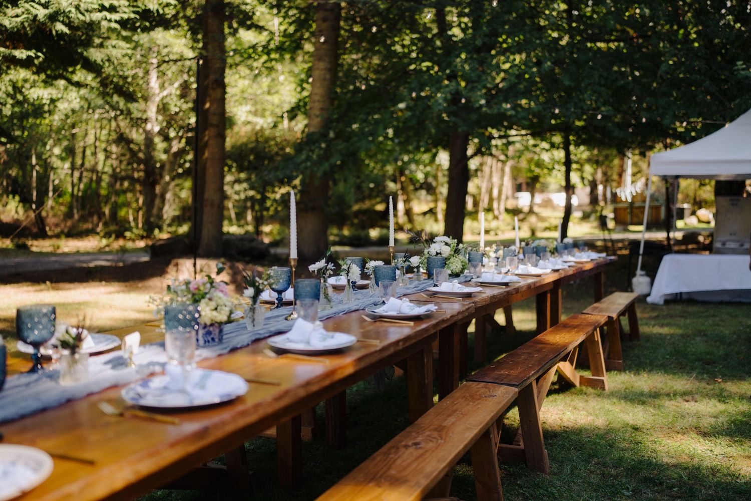 Long wooden farm tables set for an outdoor wedding reception under string lights.