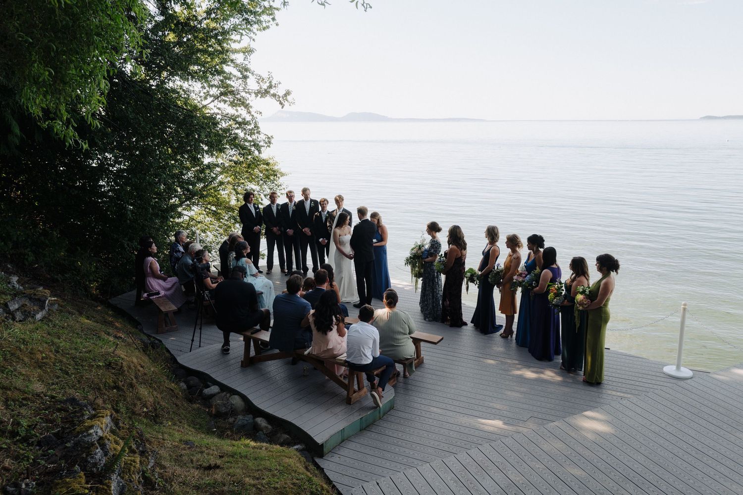 Wedding ceremony guests stand in formation on a wooden deck overlooking a serene waterfront.