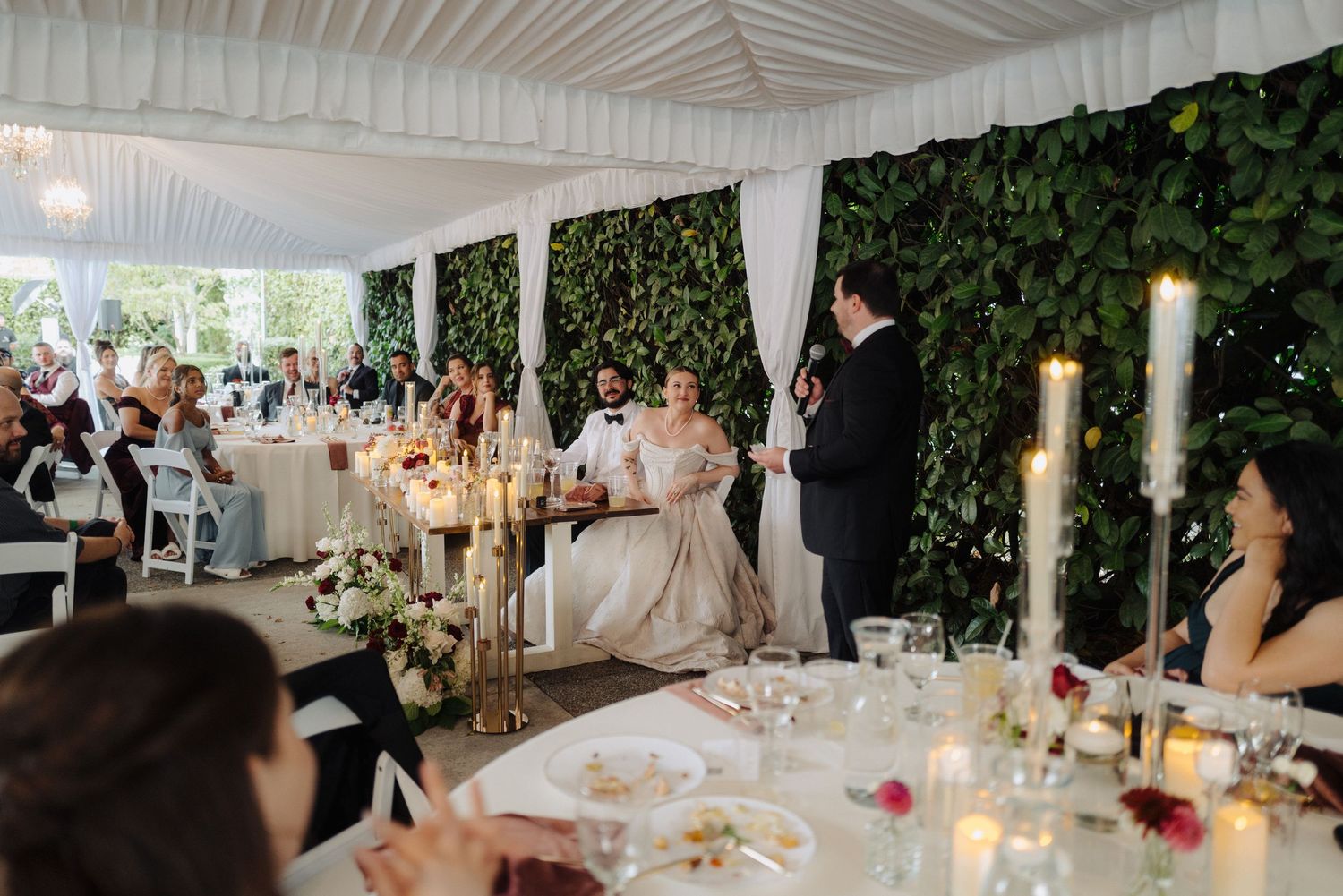 Wedding reception dining area with white draping, chandeliers and ivy-covered walls creating an intimate atmosphere.