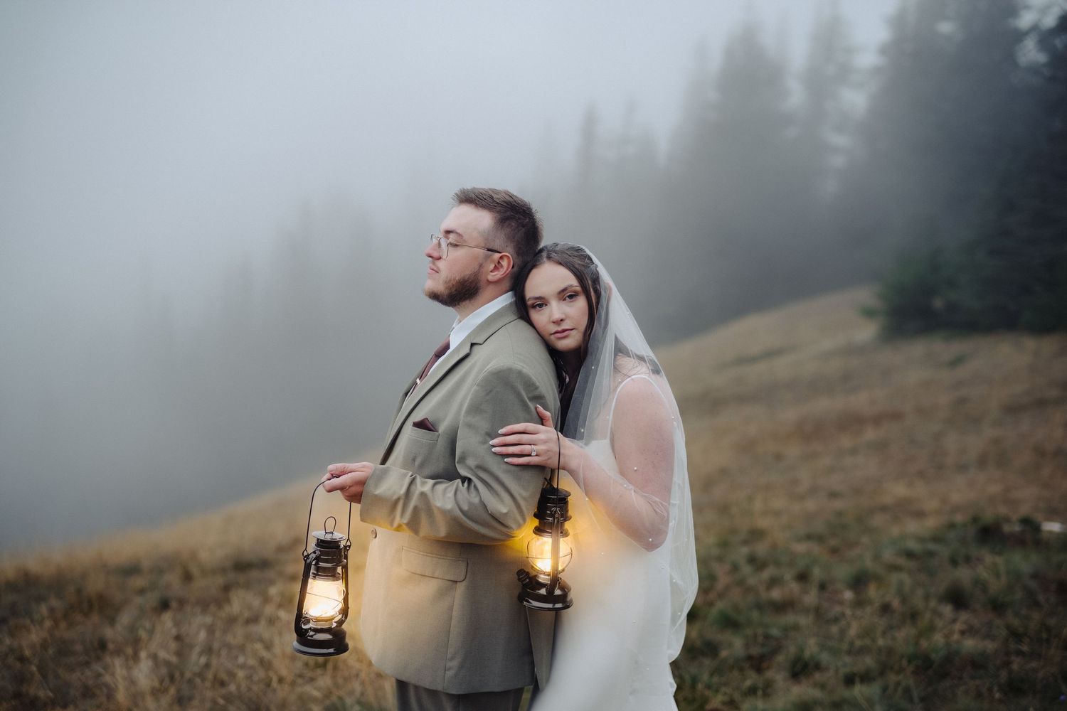 Bride and groom holding glowing lanterns back-to-back in a misty mountain meadow during their outdoor elopement.