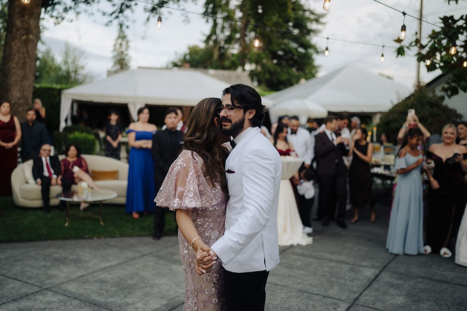 Outdoor wedding reception with guests dancing under string lights and white tents.