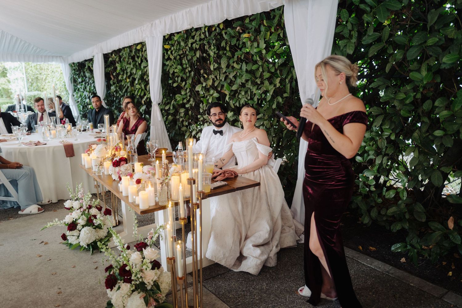 Elegant wedding reception setup with white draping, greenery walls, and glowing candle centerpieces.
