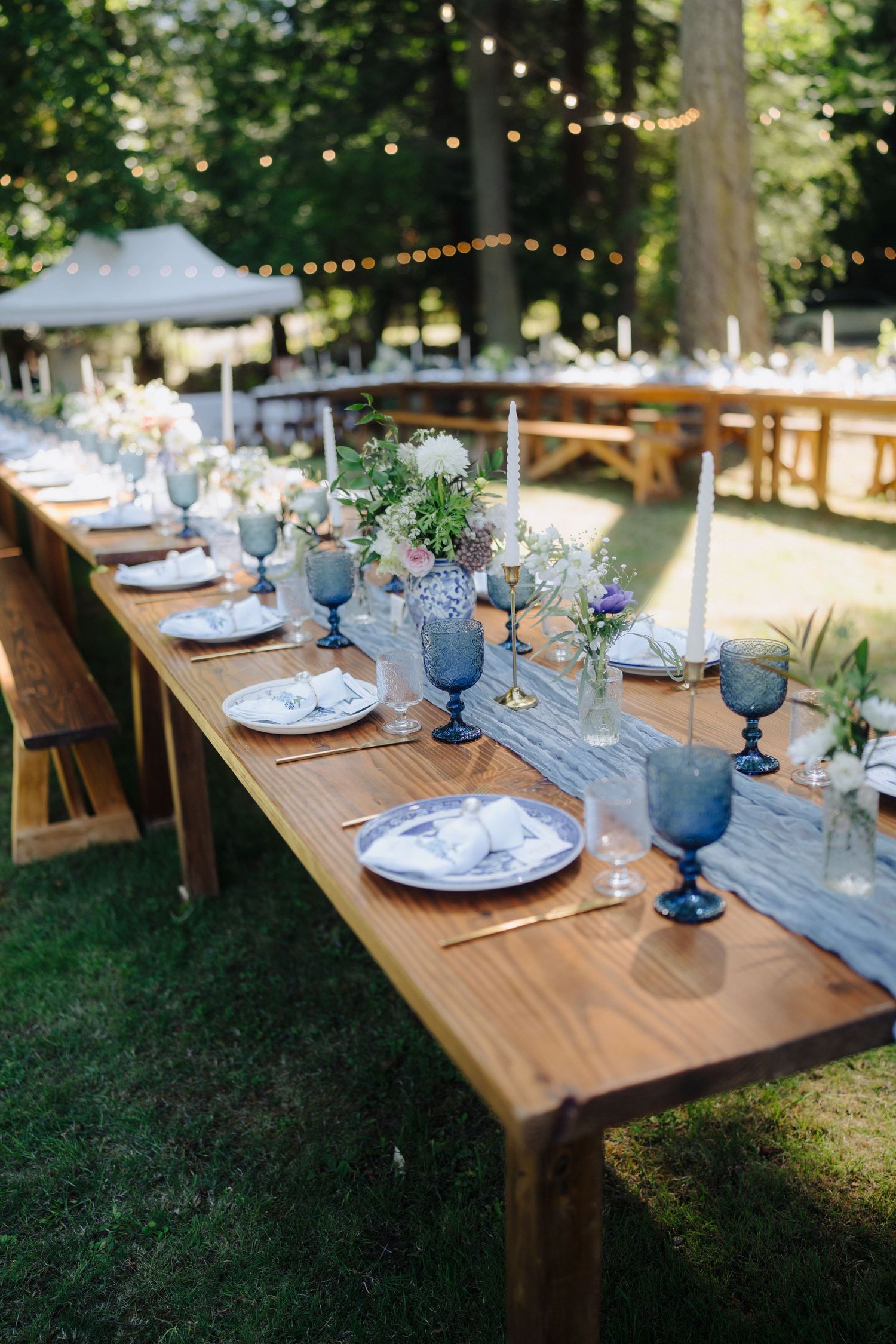 Rustic outdoor wedding reception table setting with wooden tables, blue glassware, and white place settings in a garden.