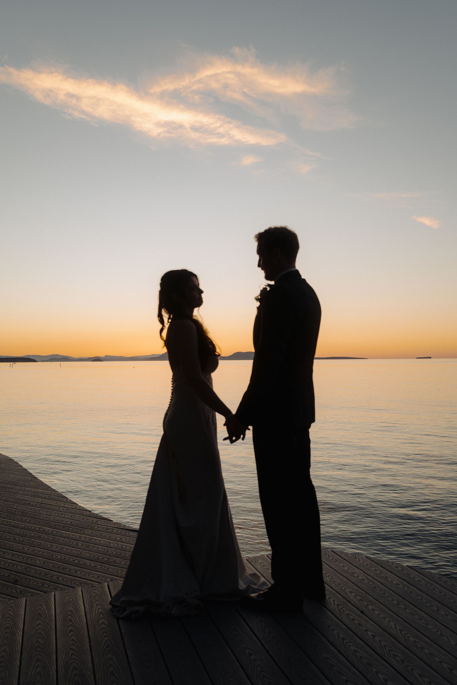 Silhouetted couple stands on wooden dock at sunset over tranquil ocean waters in romantic wedding photo series.
