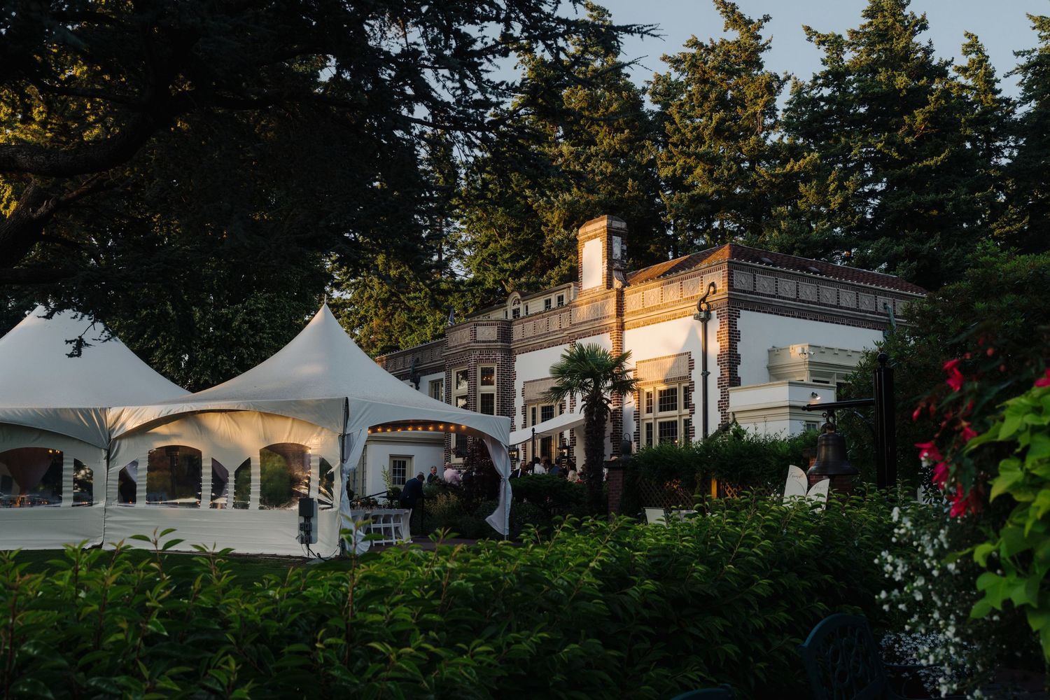 White wedding tent and elegant mansion venue surrounded by evergreen trees at sunset.